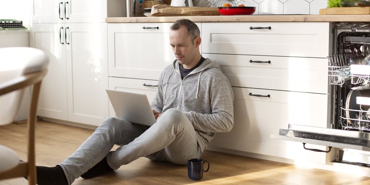 Man studying a Blended Learning module from home, sitting on the kitchen floor with a laptop and coffee mug – flexible learning on his own terms.